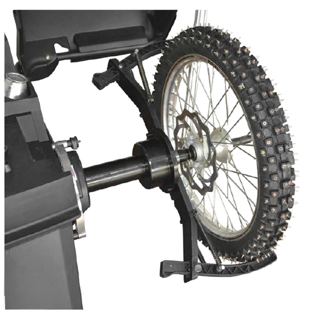 Detailansicht eines montierten Rades mit grobstolligem Spikereifen auf einer rotierenden Achse, inklusive Speichenfelge, Bremsscheibe und mechanischer Halte- und Spannvorrichtung an einer stationären Maschine.