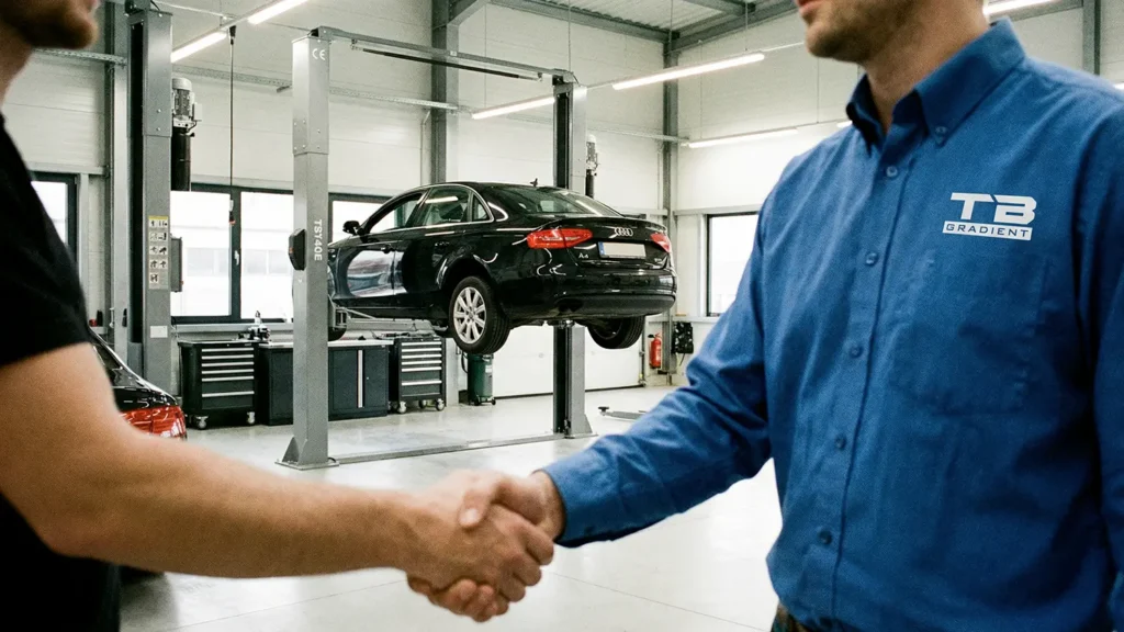 Zwei Personen geben sich in einer Werkstatt die Hand; im Hintergrund steht ein schwarzes Auto auf einer Hebebühne, rechts trägt eine Person ein Hemd mit dem Logo „TB Gradient“.